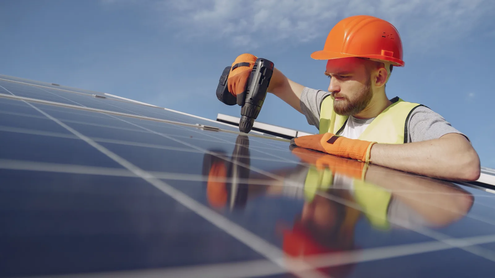 An installer in a hard hat and high-visibility vest fixing a solar panel into place against a blue sky.